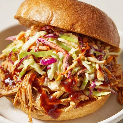 Tender pulled barbecue chicken nestled in a soft brioche bun, layered with vibrant cabbage slaw and a drizzle of sauce.  